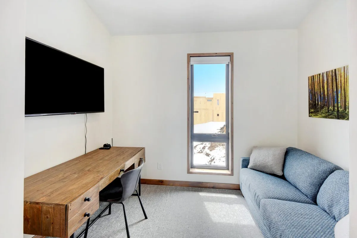 A quiet office nook with a wooden desk, sofa, and a large window offering tranquil views and natural light. Forest-themed artwork completes the serene atmosphere—perfect for remote work or planning adventures.