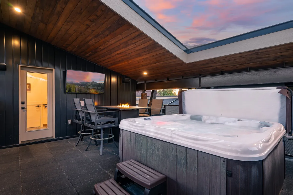 This image displays a rooftop deck featuring a hot tub, outdoor seating, a fire pit, and a wall-mounted TV. The atmosphere is tranquil and inviting, with a focus on both relaxation and entertainment, highlighted by sunset lighting.