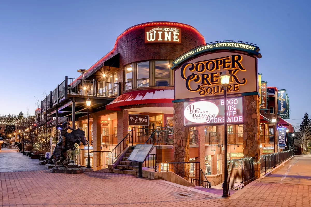 Cooper Creek Square’s vibrant exterior, featuring inviting shop signs, a staircase leading up to dining terraces, and an outdoor dining area bustling with people. The lively atmosphere hints at year-round activity, including shopping, dining, and community gatherings.