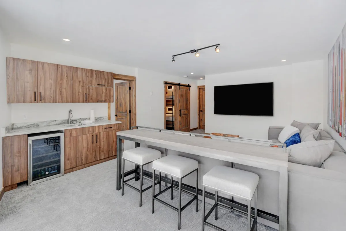 A lively, contemporary lower-level great room with a bar area, sleek kitchen cabinetry, comfortable seating, and a prominent television. The arrangement suggests both entertaining guests and relaxing movie nights. The mood is relaxed and perfect for group celebrations or après-ski evenings.