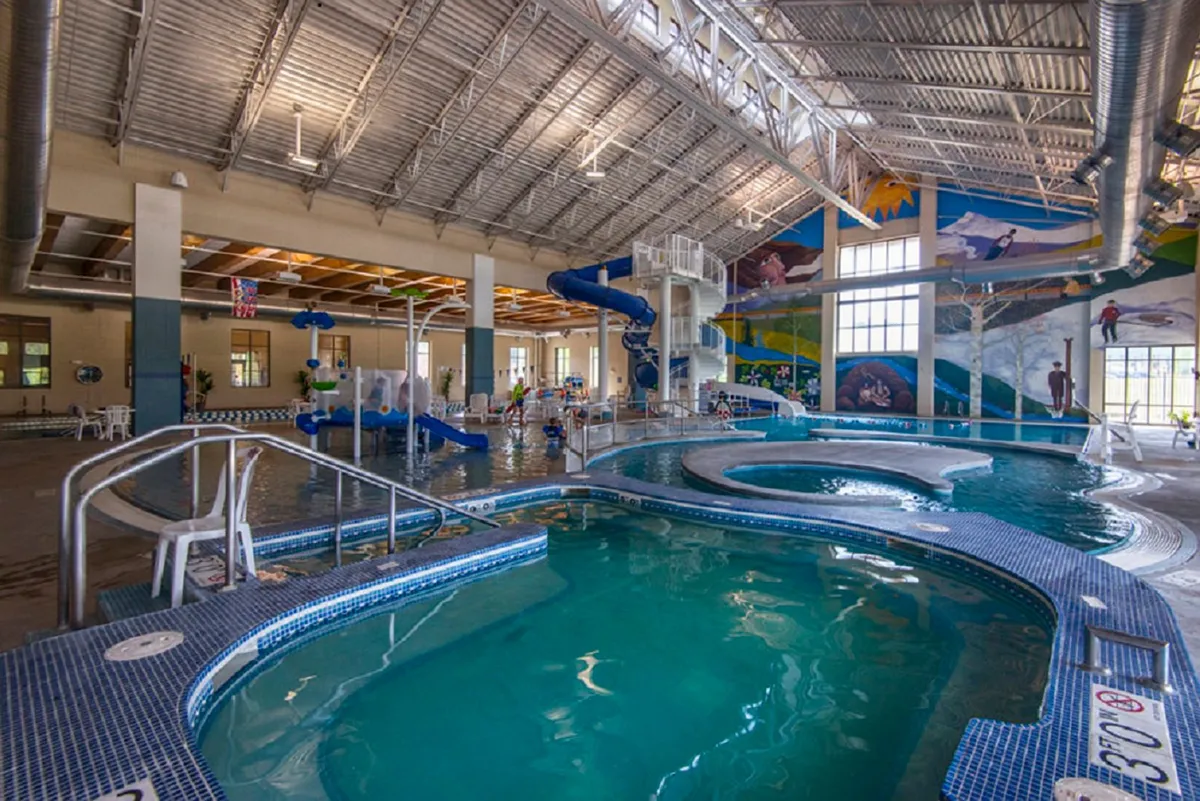 A vibrant, energetic scene at a pool facility featuring water slides, a swimming pool, colorful murals, and children joyfully playing. The atmosphere is lively and communal, evoking the excitement of family reunions and celebrations.