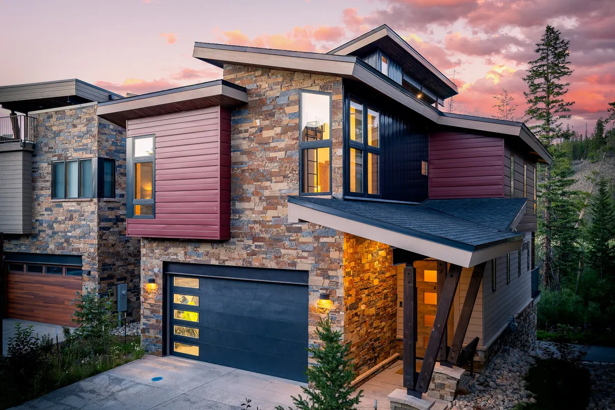 A welcoming exterior view of the home showcasing its stone facade, colorful siding, modern roof angles, and large windows. The inviting porch and surrounding landscape suggest a space ideal for family gatherings and outdoor activities.