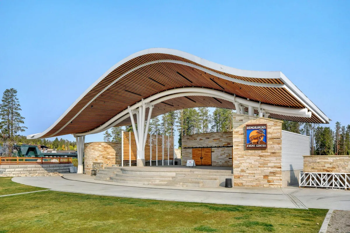 The modern, curved roof structure and stone facade of Hideaway Park, set among wooden elements and open space, creates a perfect backdrop for festivals, concerts, and community events during the warmer months.