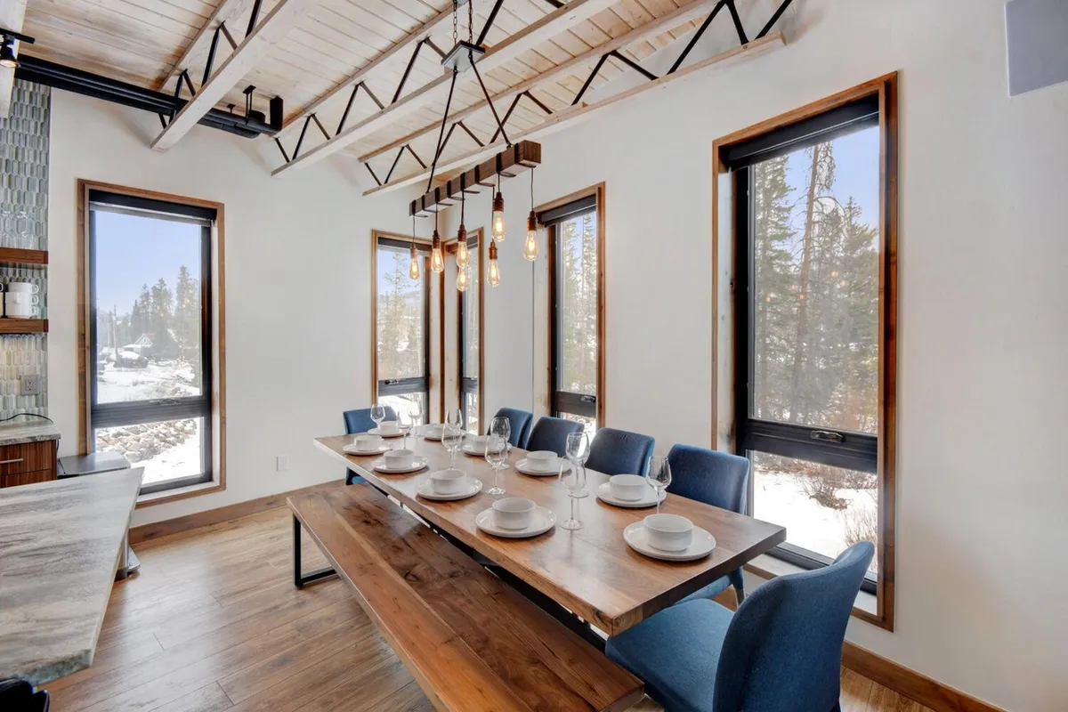 A large wooden dining table is set beneath a striking hanging light fixture, surrounded by windows that offer scenic views and flood the dining area with natural light. The setting is warm and inviting, perfect for group meals and celebrations.