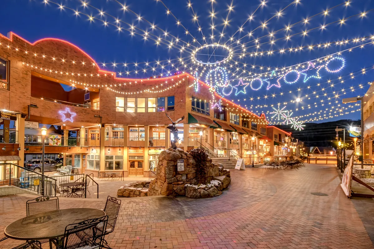 A lively downtown Winter Park setting featuring festive string lights, decorative star motifs, an outdoor seating area, and a stone fountain. People are seen dining, shopping, and enjoying social gatherings, highlighting the area’s vibrant atmosphere and community events.