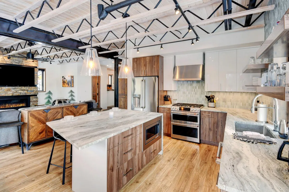 A contemporary open-concept space featuring a prominent kitchen island with sleek countertops, stainless steel appliances, an adjacent stone fireplace, and an airy layout that blends cooking, dining, and relaxation areas together.