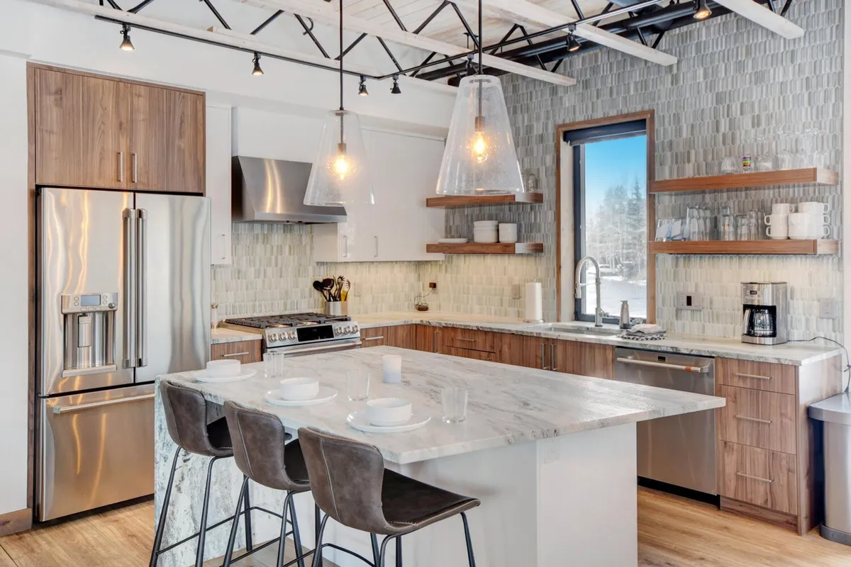 This image shows a spacious, modern kitchen featuring a large kitchen island, state-of-the-art appliances, stylish pendant lights, and ample windows letting in natural light with views of the outdoors. The space is clean and inviting, ideal for preparing gourmet meals or gathering with friends and family.
