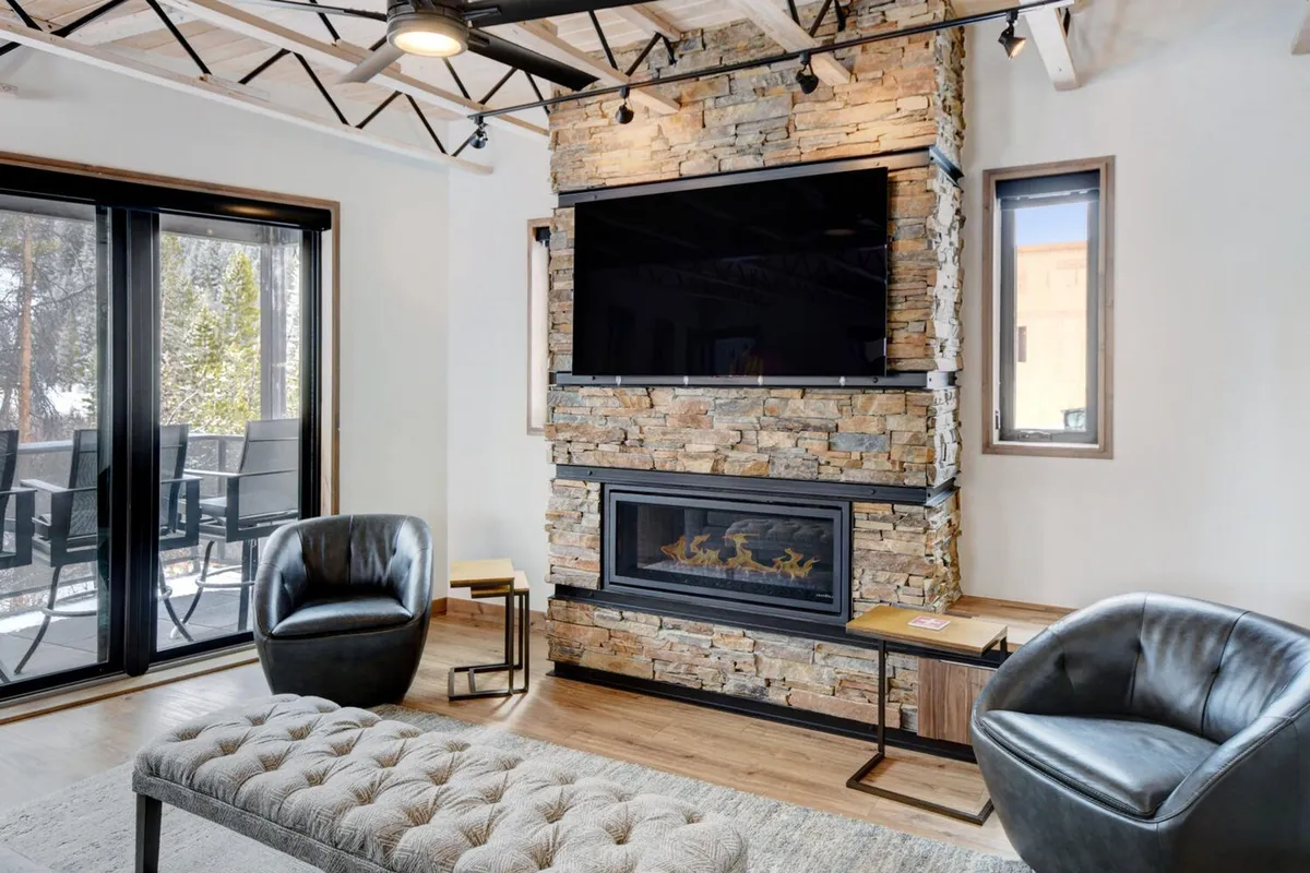 A cozy and modern great room featuring a large stone fireplace as the centerpiece, flanked by comfortable seating and a spacious area for socializing. A wall-mounted TV is visible above the fireplace, and expansive windows frame beautiful outdoor views, bathing the space in natural light.