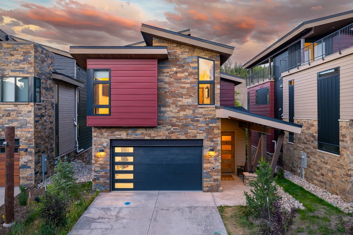 A contemporary chalet exterior featuring modern architecture, a stone facade, colorful siding, and a landscaped entryway. In the foreground, family members gather and relax on the porch as the sun sets, conveying a welcoming atmosphere that hints at special occasions and social gatherings.